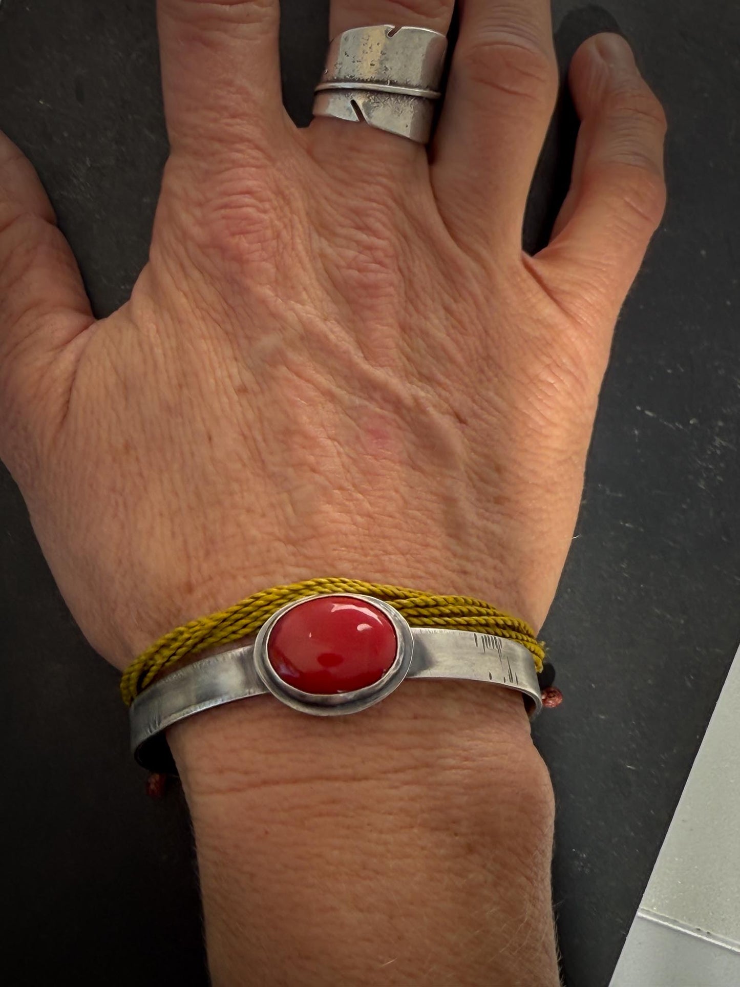 Hand wearing a silver bracelet with a red stone and yellow cord on a dark background