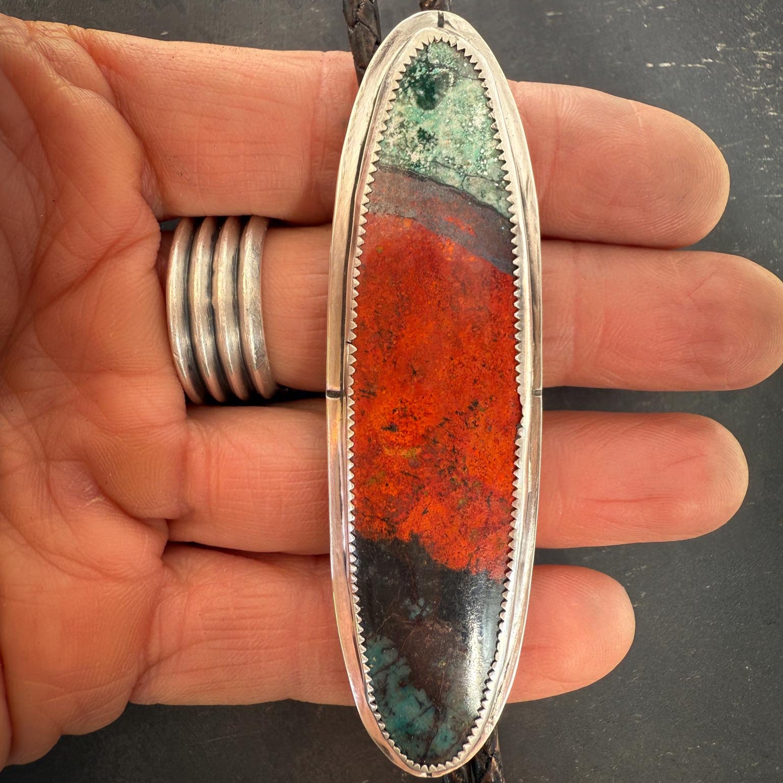 sonoran sunrise bolo tie held in a hand with silver rings on a dark background