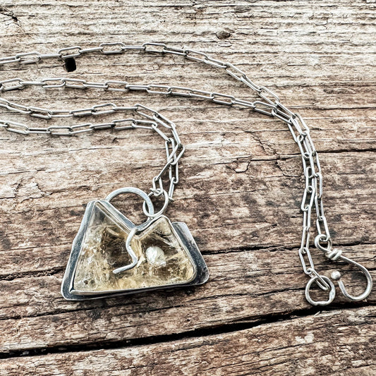 carved citrine stone, shaped like a mountain with two peaks. The stone is set in a sterling silver bezel with a singular prong, which mimics a river flowing between the peaks. The back of the pendant is cut out, creating space for the light to shine through. It hangs from a delicate paperclip chain, and is oxidized silver to complement the warm tones of the citrine.