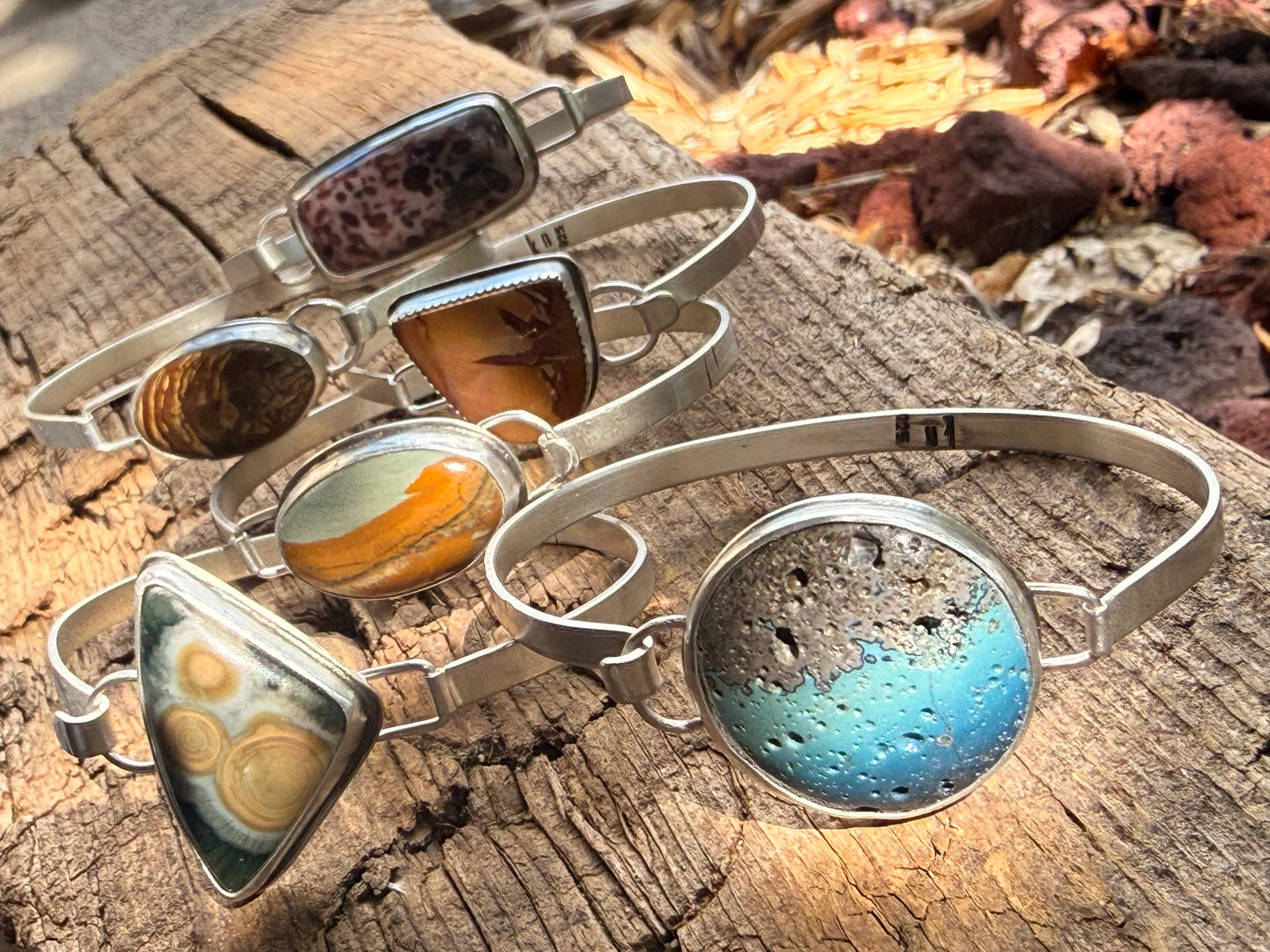 Five silver bangle bracelets with various colorful natural stone cabochons, displayed on a rustic wooden log outdoors.