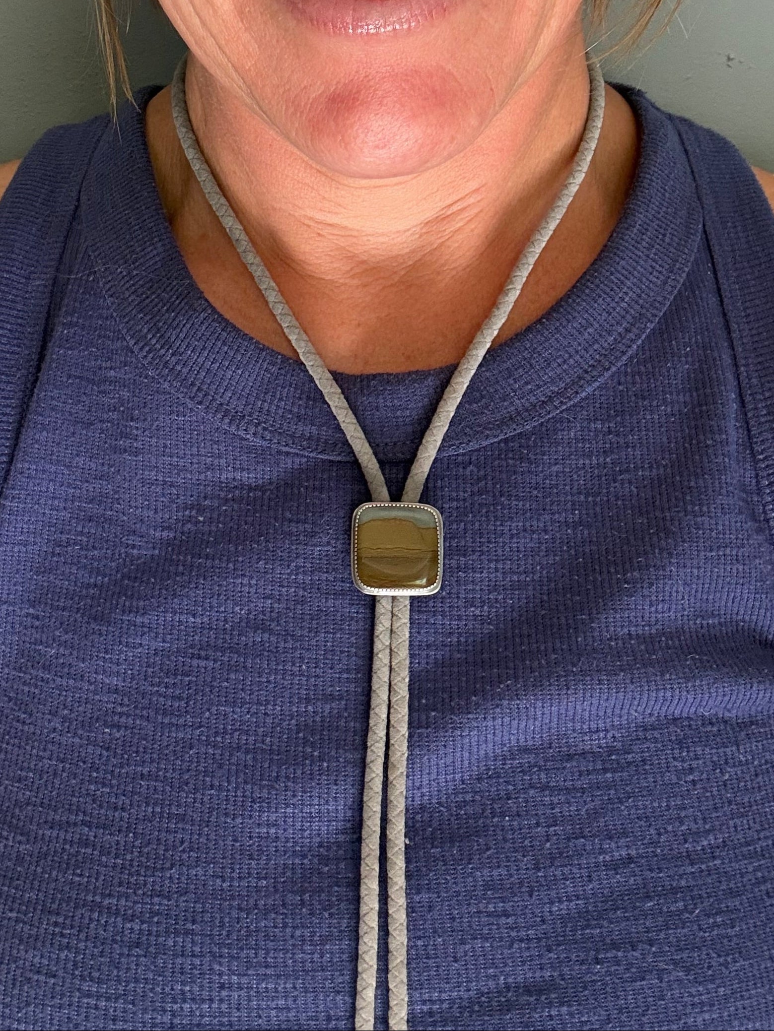 A close-up shot of a woman wearing a gray braided bolo tie necklace with a square metallic pendant, paired with a purple ribbed tank top.
