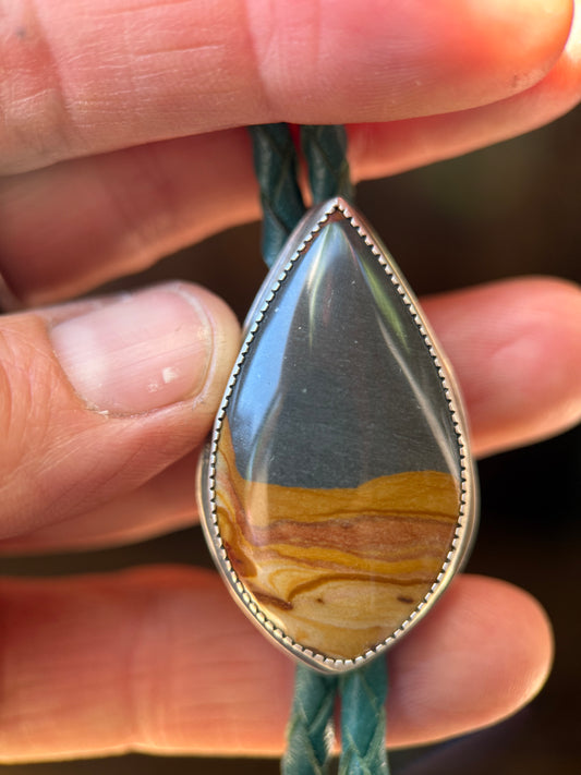 A close-up of a hand holding a silver bezel pendant necklace with a teardrop-shaped natural stone showing earthy yellow, brown, and gray banding, suspended on a dark green braided cord.