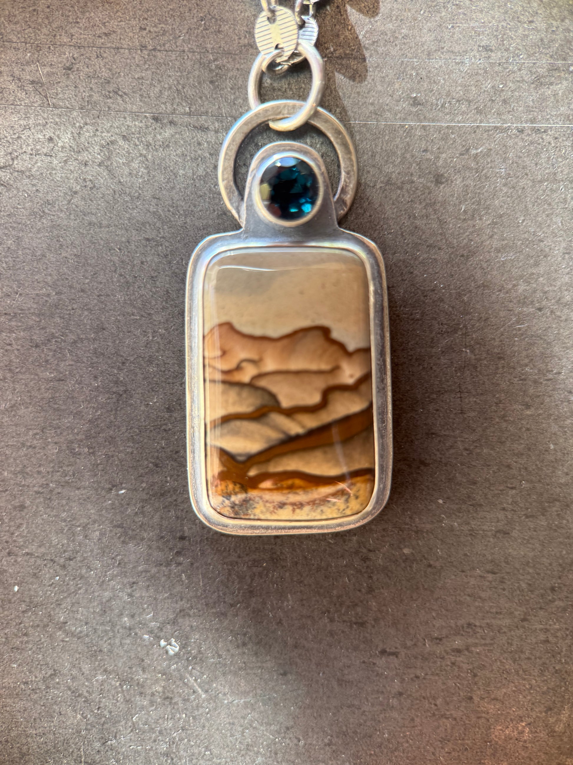 A silver-toned rectangular pendant necklace featuring a natural agate or jasper landscape inlay with tan, brown, and cream bands, accented by a small round dark blue gemstone at the top.