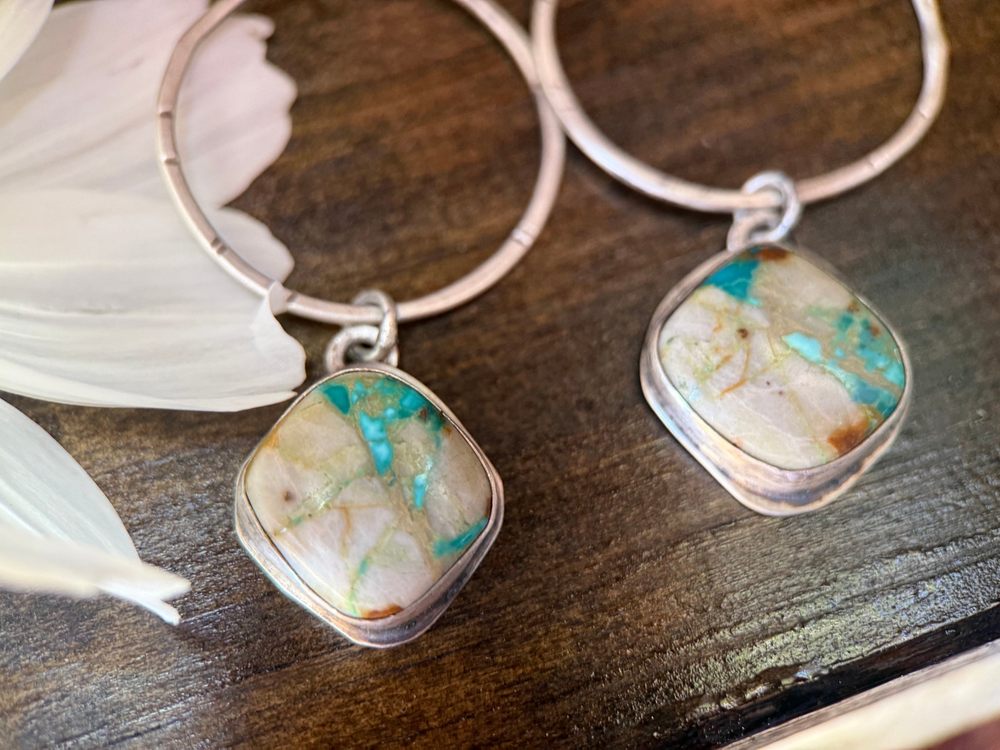 A pair of silver hoop earrings with square bezel-set turquoise and cream marbled stone pendants, displayed on a dark wooden surface next to white feathers.