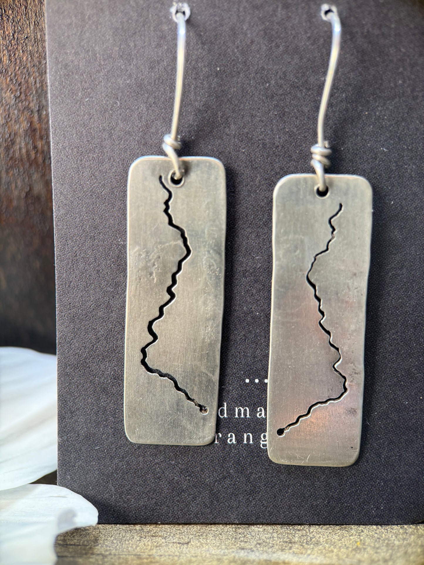 A pair of handmade silver drop earrings with rectangular metal plates featuring a jagged crack design, hanging from silver fishhook wires on a dark display card.