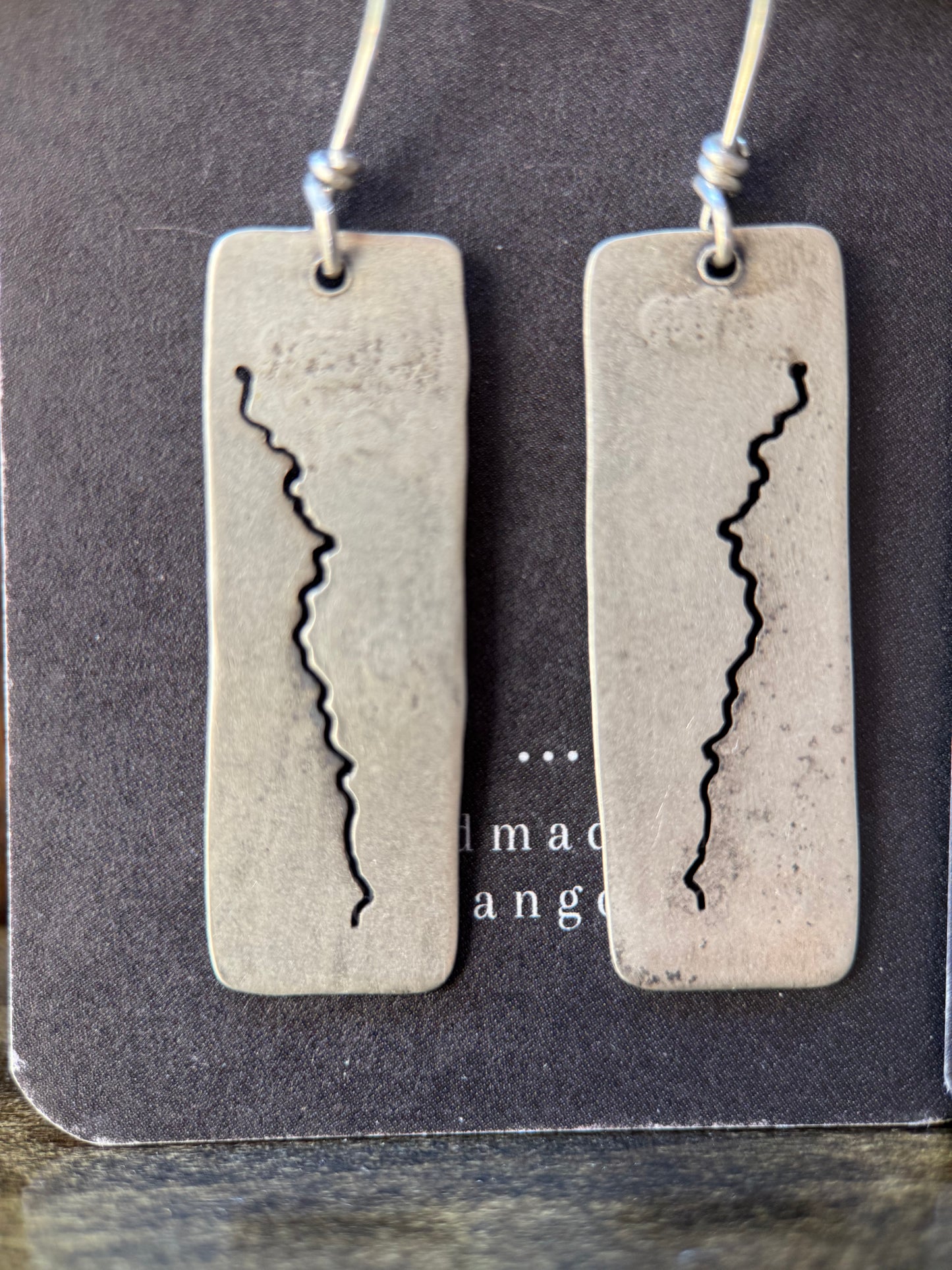 A pair of rectangular silver-toned earrings with a jagged vertical crack design, hanging from thin cords on a dark display card.