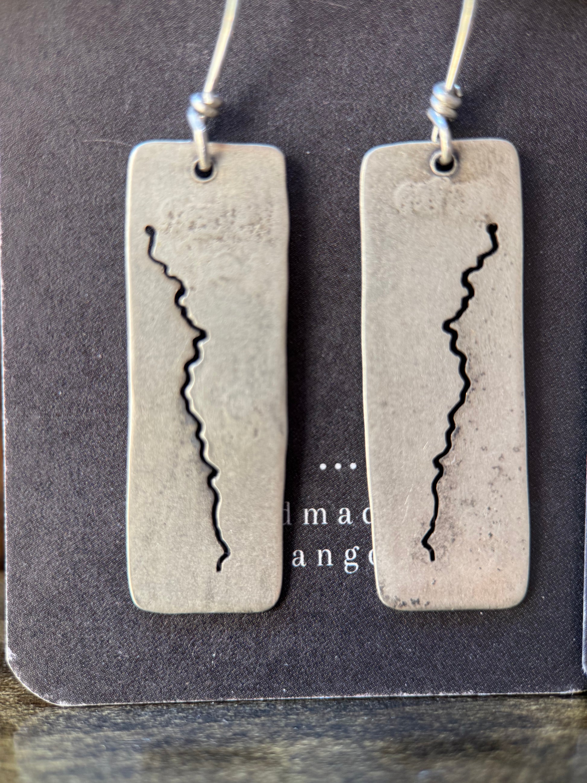 A pair of rectangular silver-toned earrings with a jagged vertical crack design, hanging from thin cords on a dark display card.