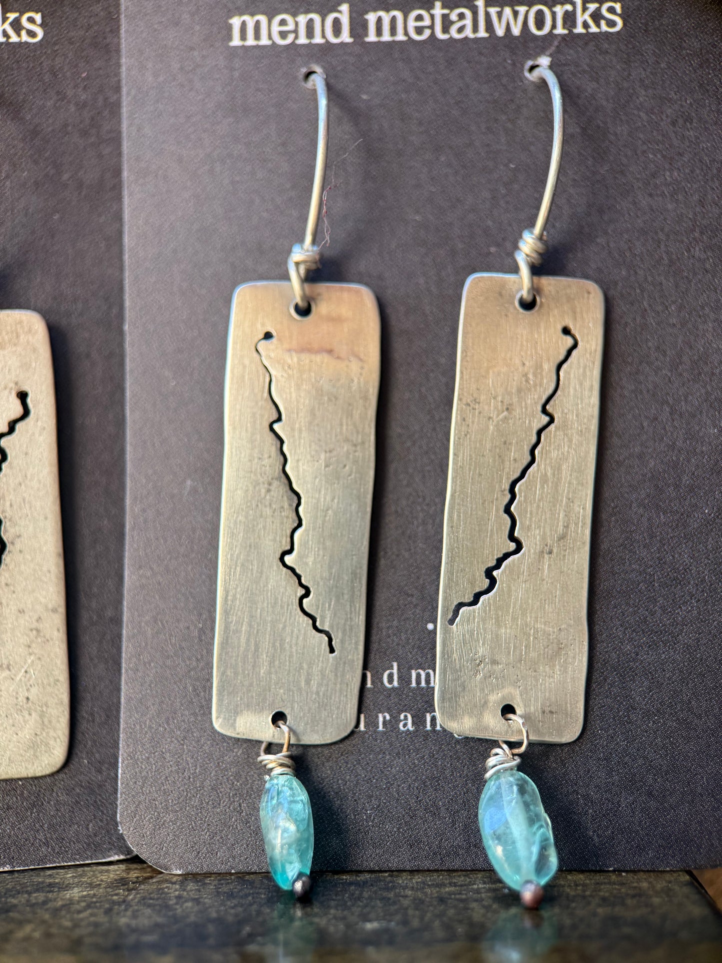 A pair of handmade dangle earrings with rectangular brushed silver-tone metal plates featuring a jagged cutout design, hanging from silver hooks with blue faceted beads below, displayed on a dark card.