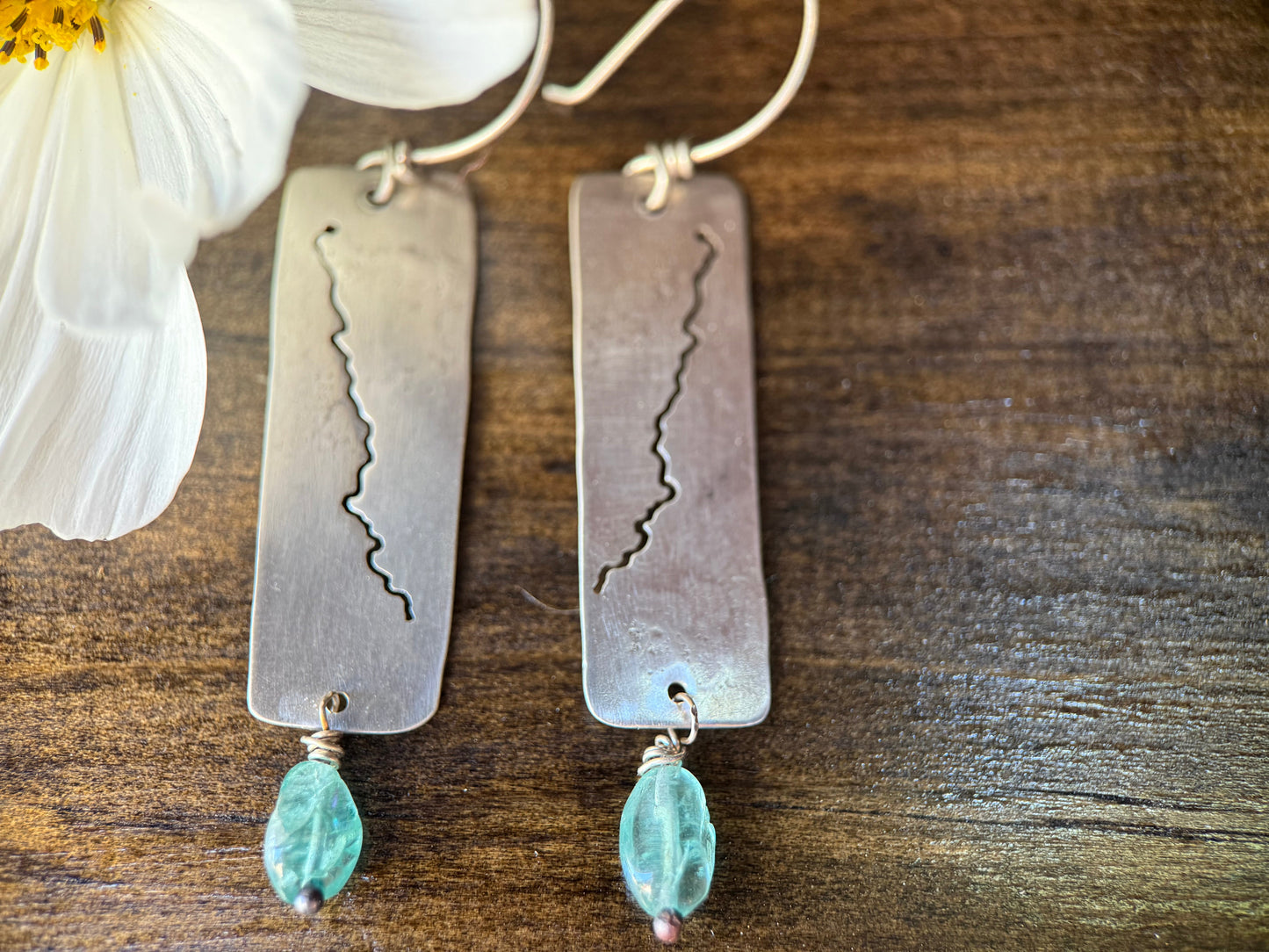 A pair of handmade silver dangle earrings with rectangular metal plates featuring an engraved mountain range pattern, accented with small light blue beads, displayed on a dark wood surface next to a white flower.