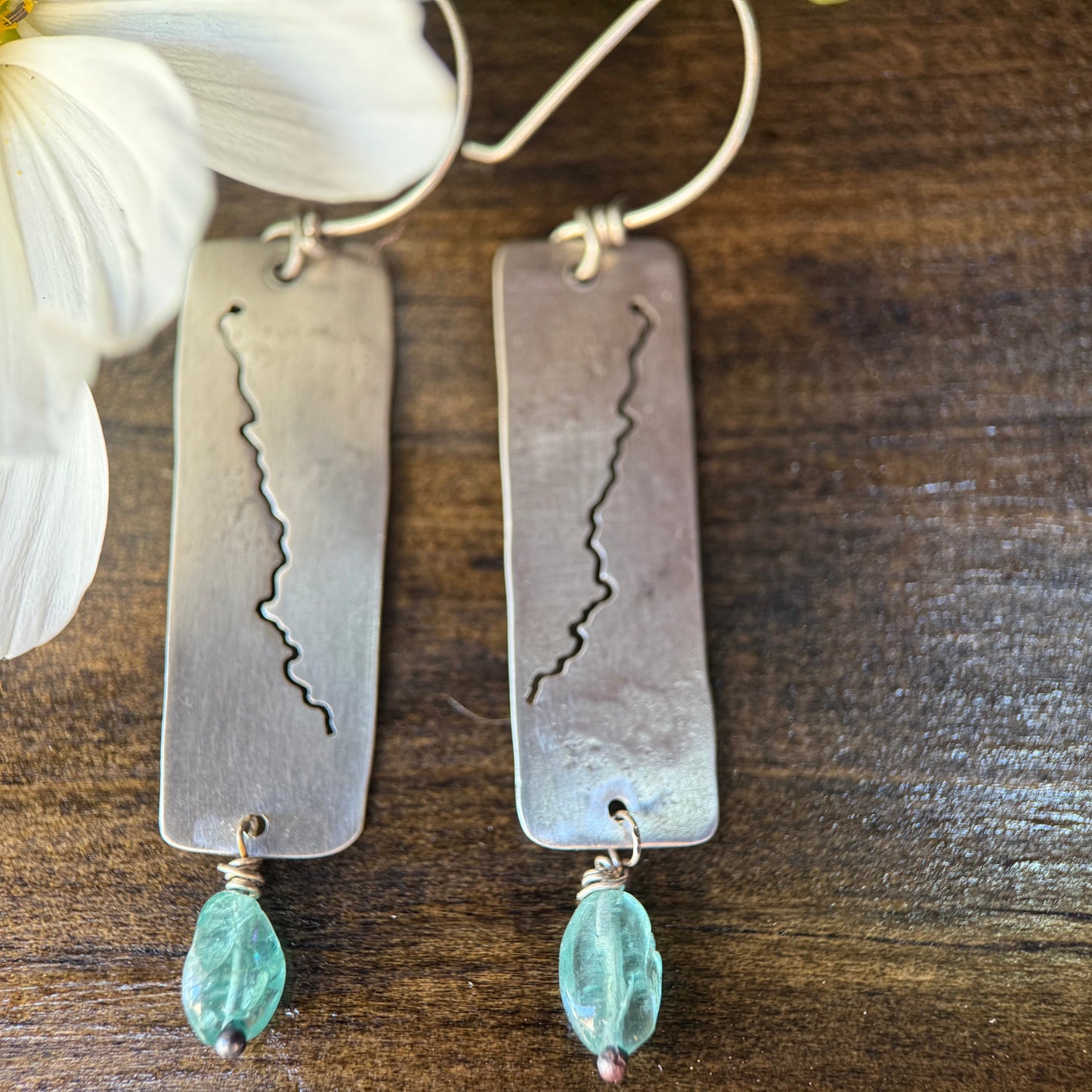 A pair of handmade silver drop earrings with rectangular plates featuring a hand-drawn mountain range line design, hanging from silver hoops and accented with small light blue faceted beads, displayed on a dark wood surface next to a white flower.