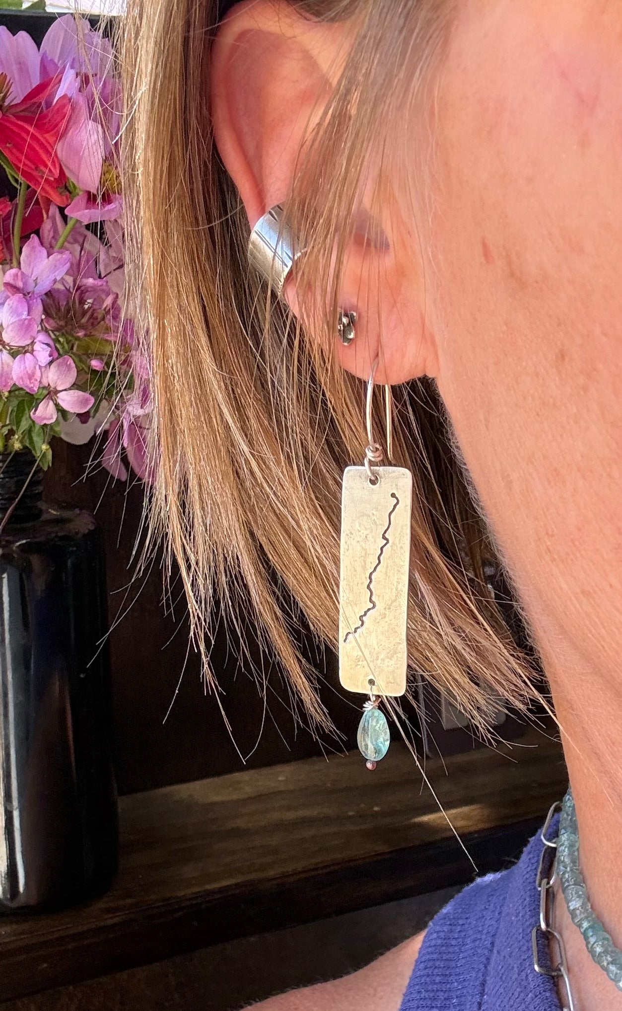 A close-up of a person's ear wearing a long rectangular drop earring made from a natural wood slice with a black lightning-bolt design, hanging from silver rings, with a small light blue bead dangling at the bottom.