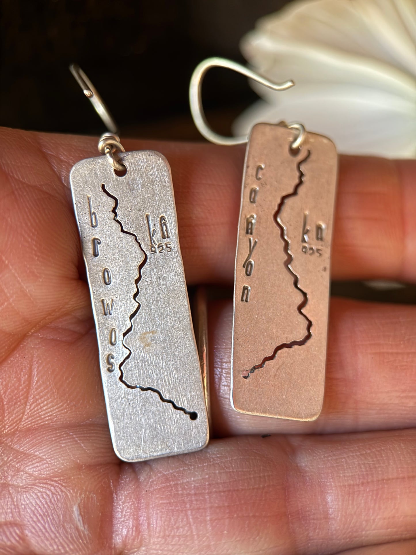 A pair of rectangular drop earrings held in a person's hand. One earring is silver-toned with a dark engraved line and the word 'browns,' and the other is a warm rose-gold or copper-toned version with a similar engraved design and the word 'cayon.