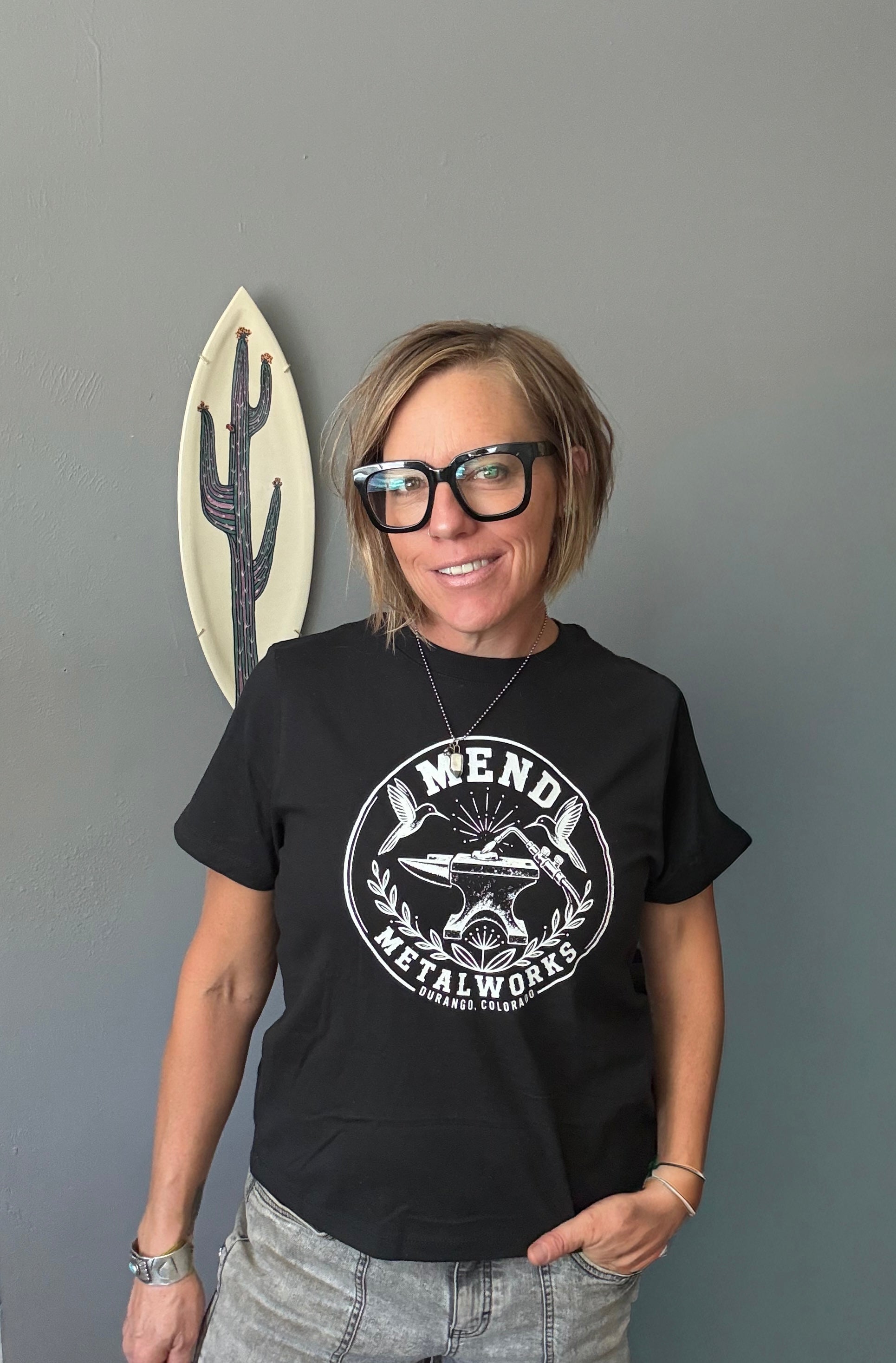 A woman with short blonde hair and black glasses wearing a black graphic t-shirt with a white circular 'Mend Metalworks' logo, standing against a gray wall