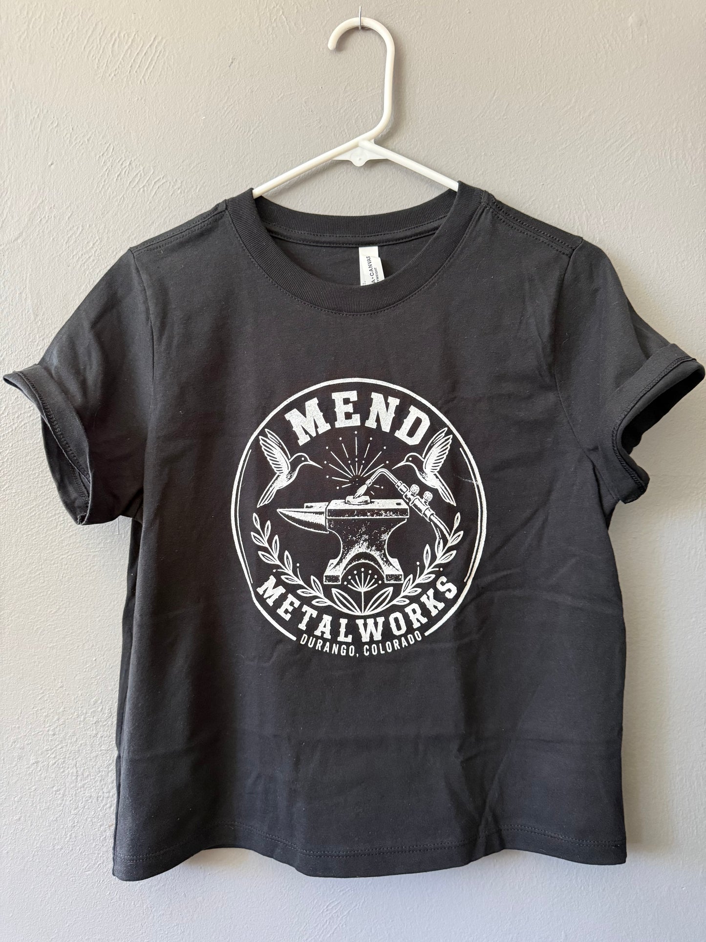 A dark charcoal gray t-shirt hanging on a white plastic hanger against a textured beige wall. The shirt features a large white circular graphic on the front with the text 'MEND METALWORKS' and an illustration of an anvil, torch, two hummingbirds and flower.
