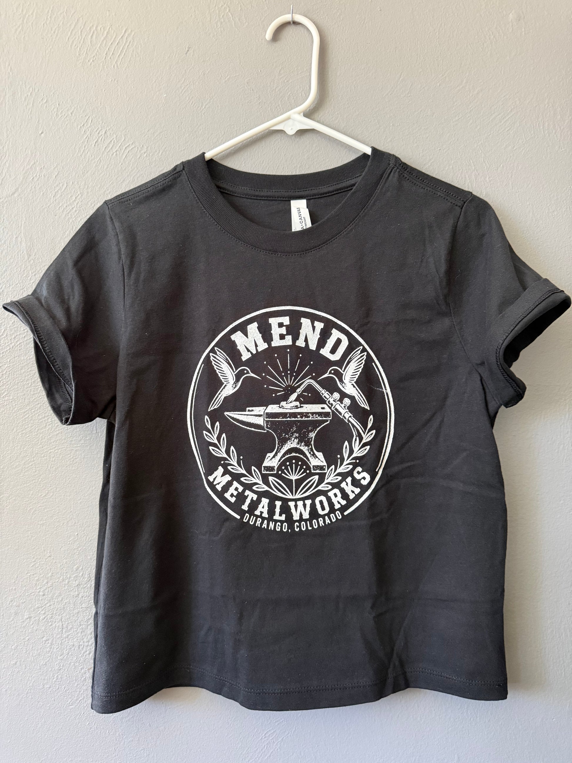 A dark charcoal gray t-shirt hanging on a white plastic hanger against a textured beige wall. The shirt features a large white circular graphic on the front with the text 'MEND METALWORKS' and an illustration of an anvil, torch, two hummingbirds and flower.