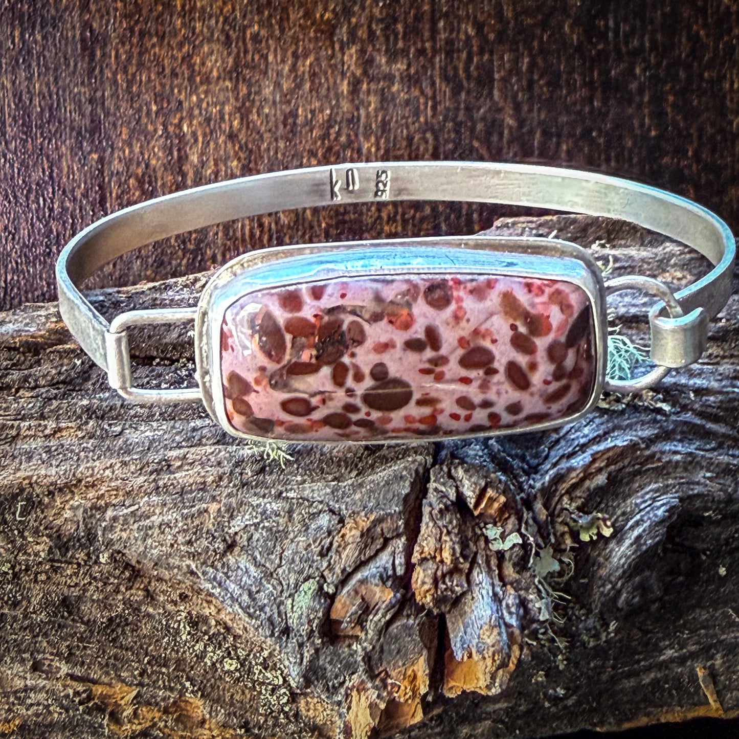 A silver-toned metal cuff bracelet with a rectangular bezel holding a pink and brown marbled stone, displayed on a piece of weathered wood against a dark wood background.