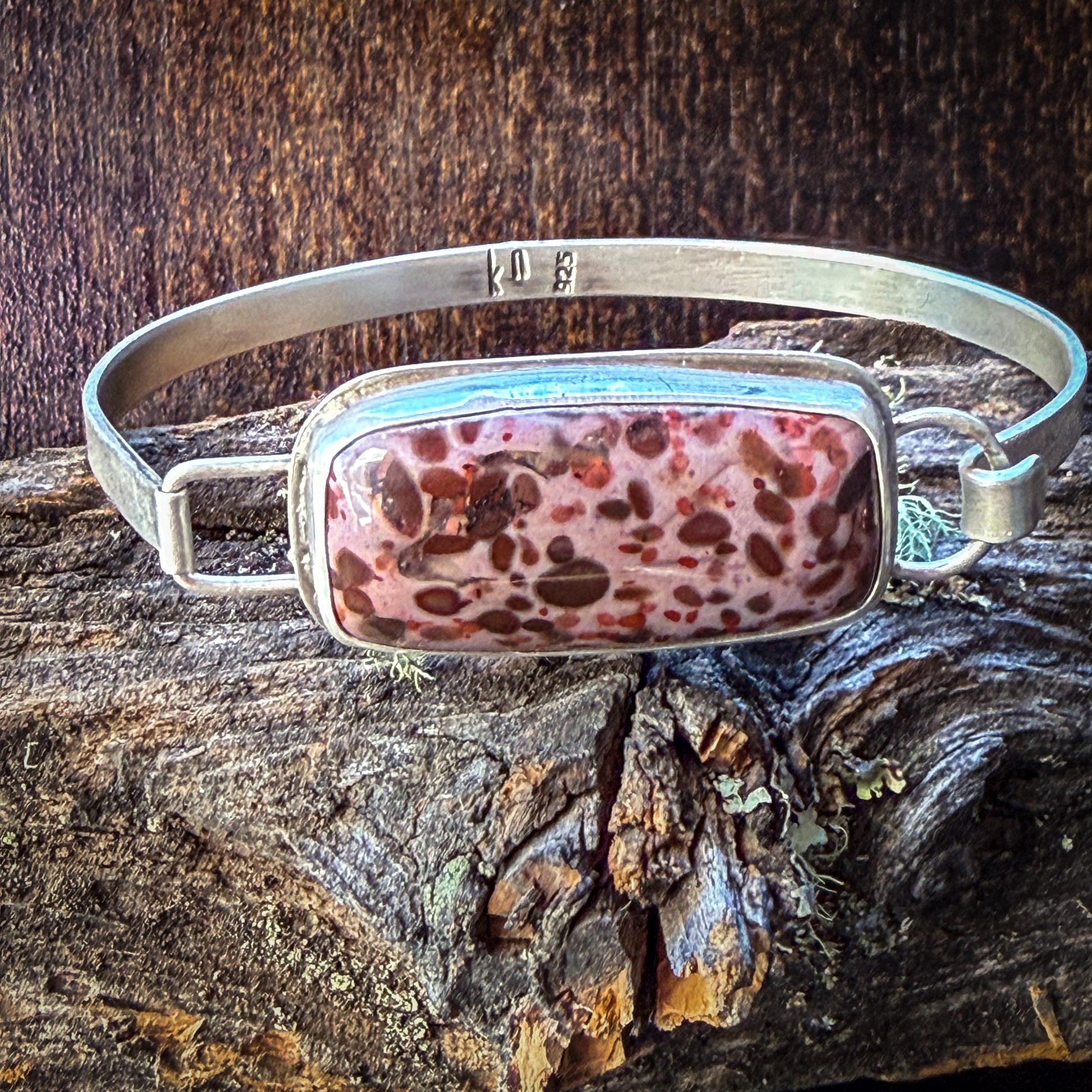 A silver-toned metal cuff bracelet with a rectangular bezel holding a pink and brown marbled stone, displayed on a piece of weathered wood against a dark wood background.