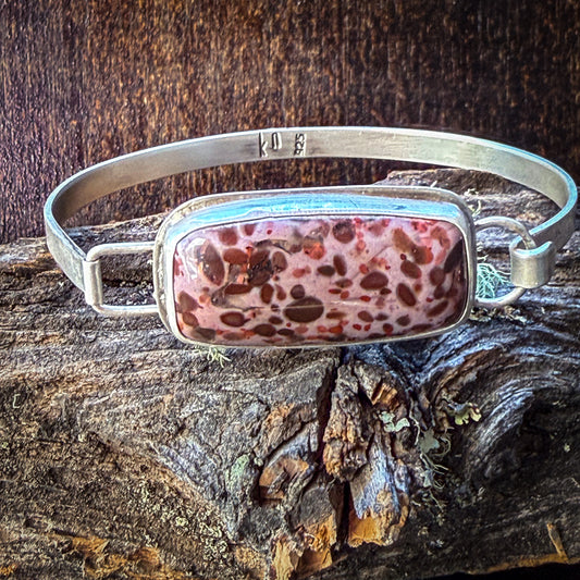 A silver-toned metal cuff bracelet with a rectangular bezel holding a pink and brown marbled stone, displayed on a piece of weathered wood against a dark wood background.