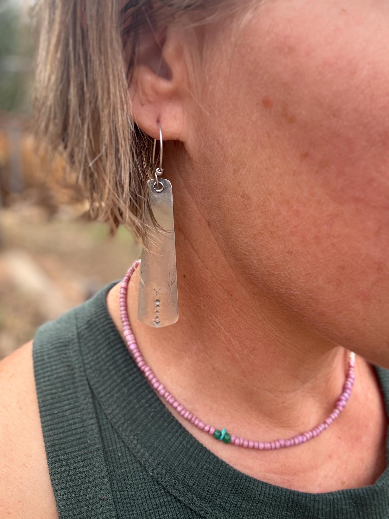 Person wearing a silver earring and pink beaded necklace with a blurred natural background