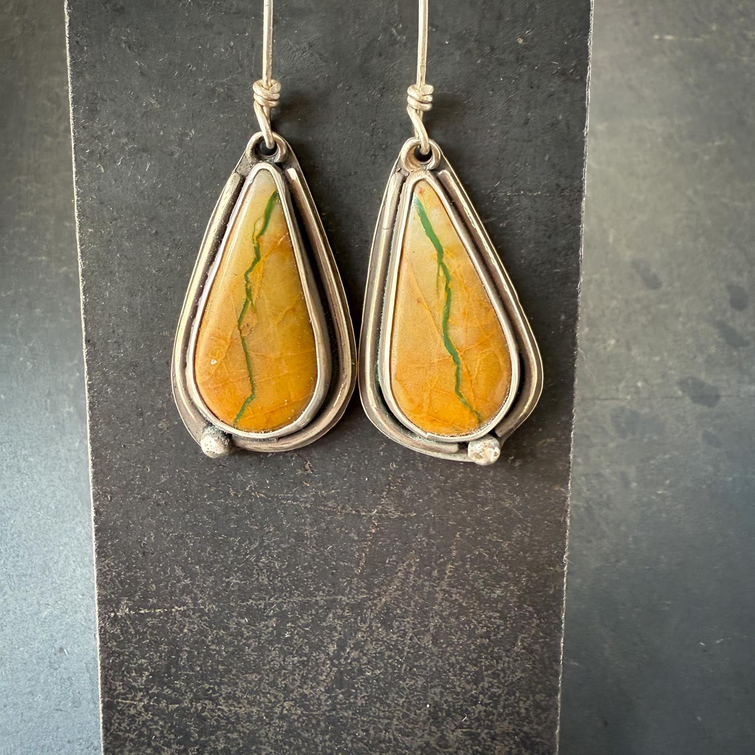 Teardrop-shaped roystone turquoise earrings with yellowish stone and matrix, with a dark turquoise vein running top to bottom, stones on a dark gray stand.