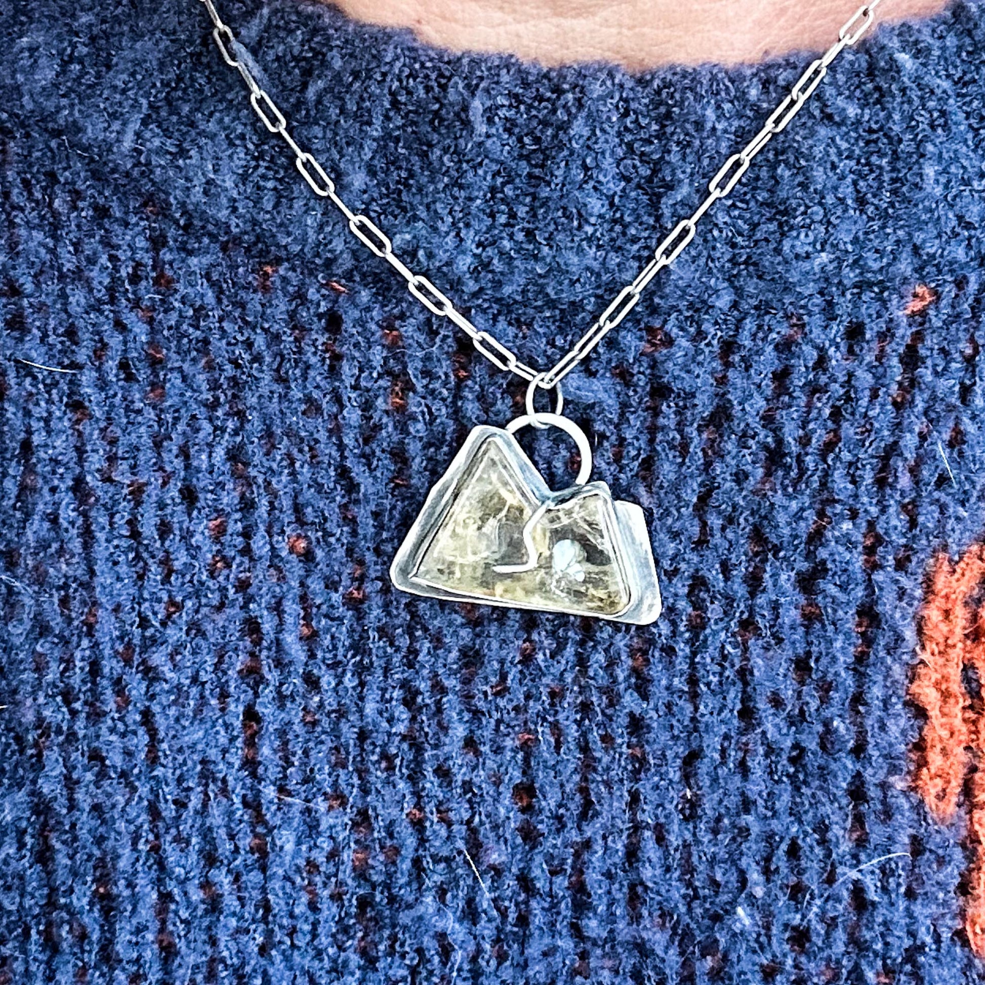 carved citrine stone, shaped like a mountain with two peaks. The stone is set in a sterling silver bezel with a singular prong, which mimics a river flowing between the peaks. The back of the pendant is cut out, creating space for the light to shine through. It hangs from a delicate paperclip chain, and is oxidized silver to complement the warm tones of the citrine.