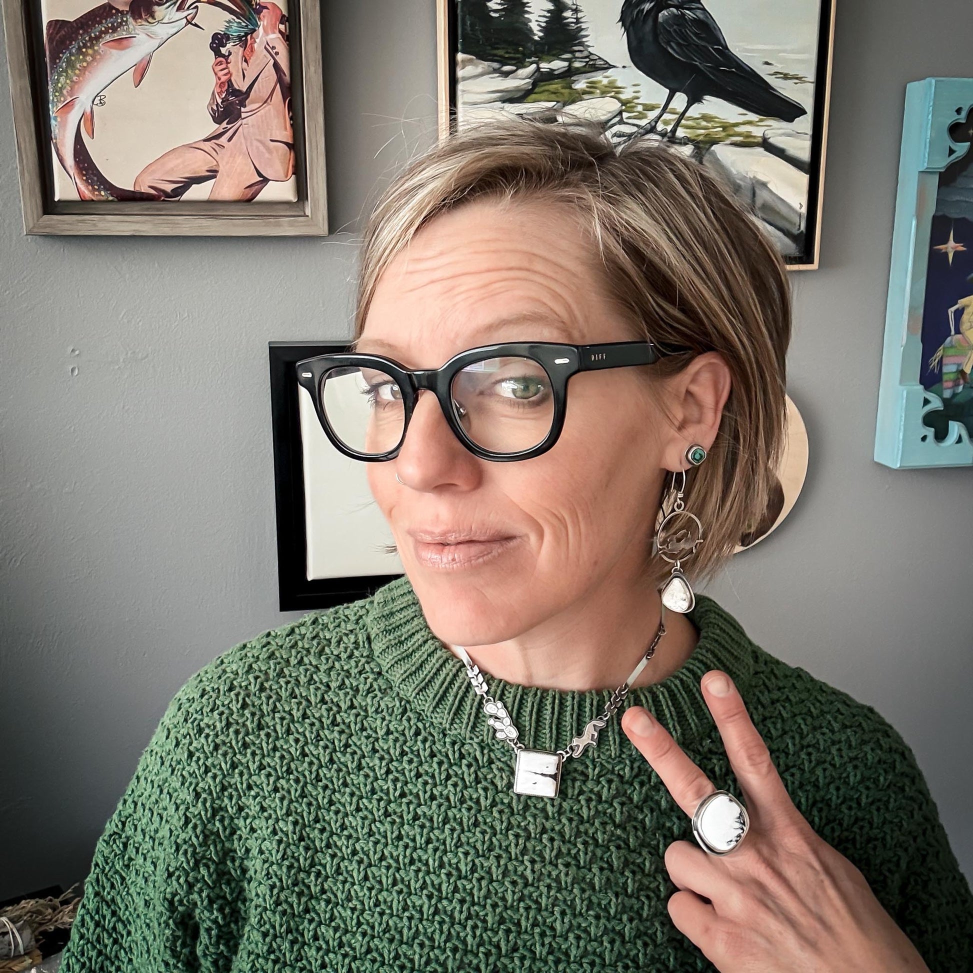 Person wearing glasses and a green sweater with framed pictures on the wall.