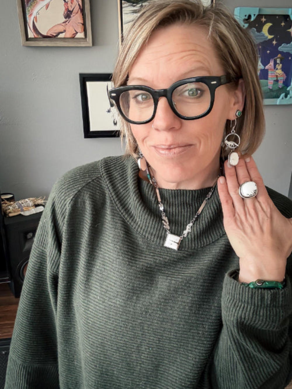 Woman wearing glasses and a green sweater with jewelry, in a room with framed pictures on the wall.