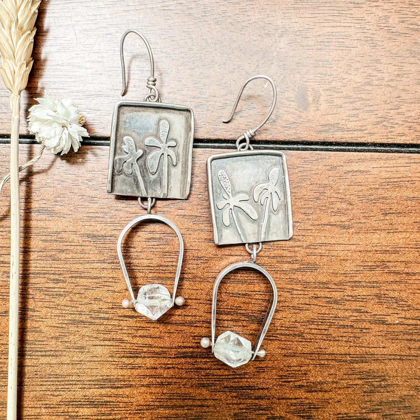 A pair of silver earrings with hand-sawn Mexican hat botanicals soldered onto a back plate. Each earring has a 3-inch drop length, a 3/4-inch ear wire, and a 1-inch horseshoe-shaped rectangle wire attached and swinging from the frame. At the horseshoe's bottom, a 3/8-inch-long faceted quartz bead is pinned in place, adding a touch of sparkle to the handcrafted design.