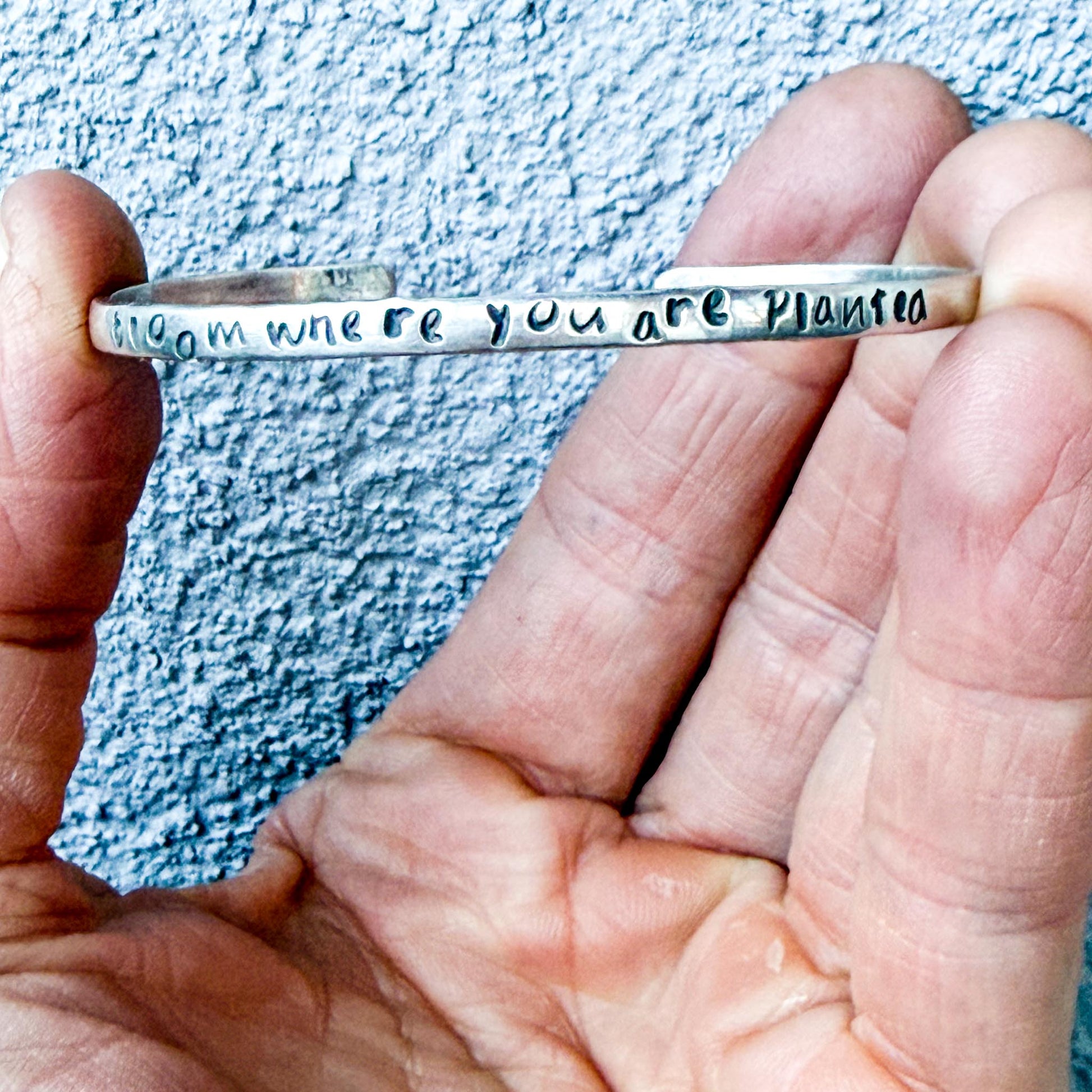 Silver bracelet with engraved text "bloom where you are planted" held by a hand against a textured blue background