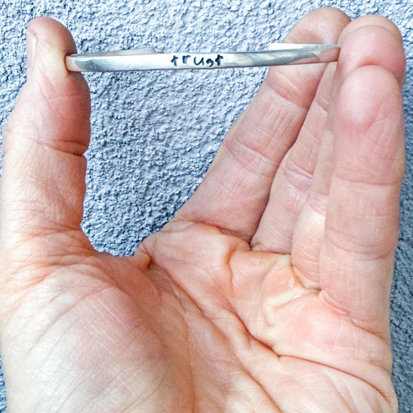 Hand holding a small silver bracelet with 'Trust' engraved on a textured blue background