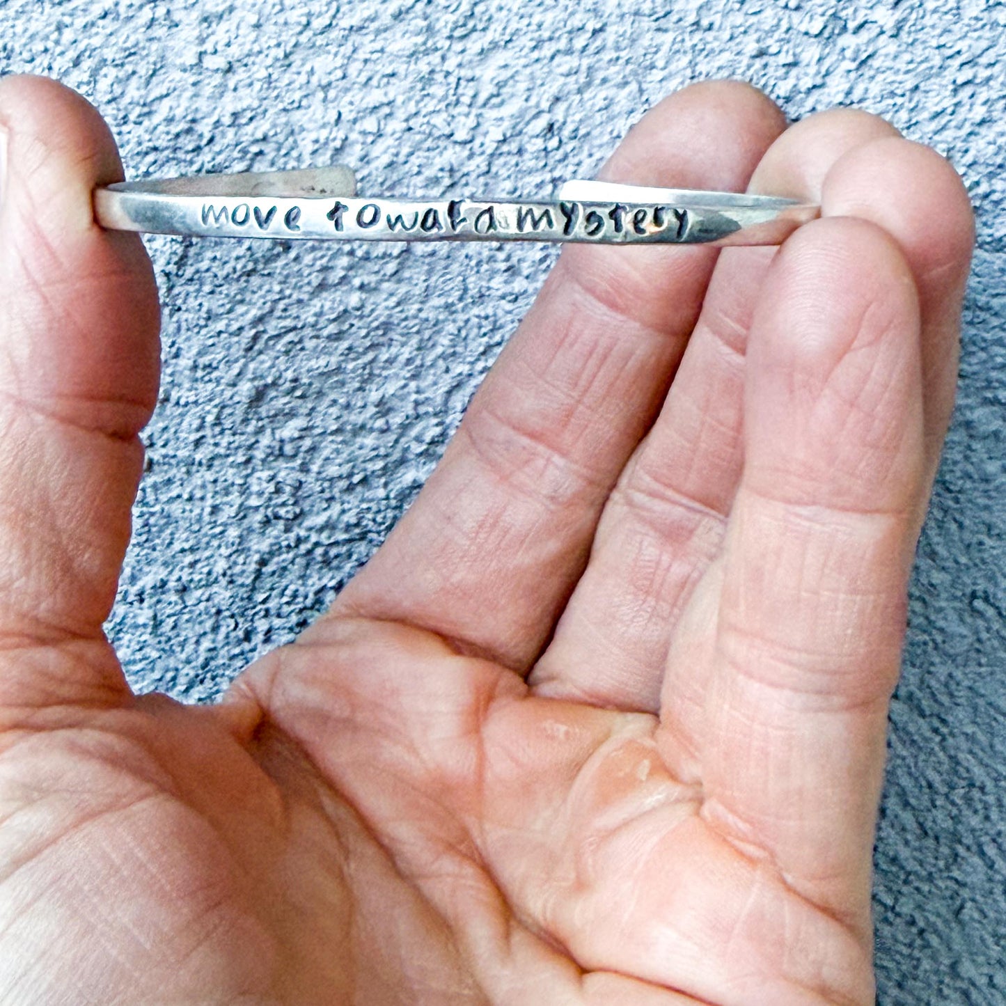 Hand holding a silver bracelet with engraved text "move toward mystery" against a textured blue background