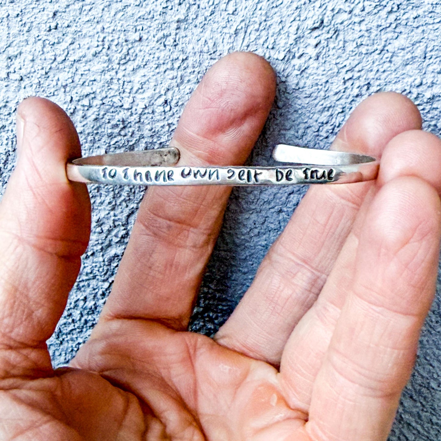 Hand holding a silver bracelet with engraved text "to thine own self be true" against a textured blue background