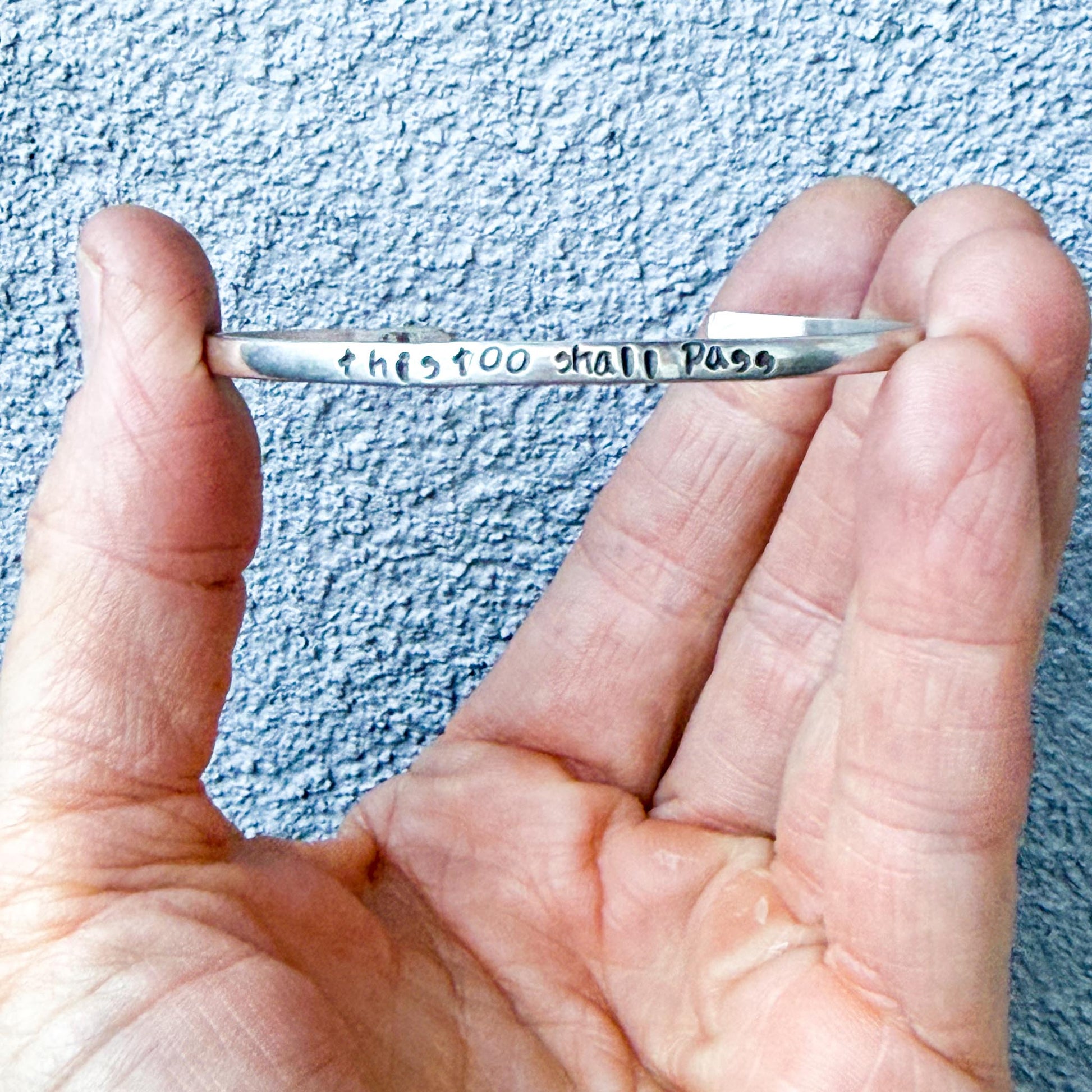 Silver bracelet with engraved text "this too shall pass" held in a hand against a textured blue background