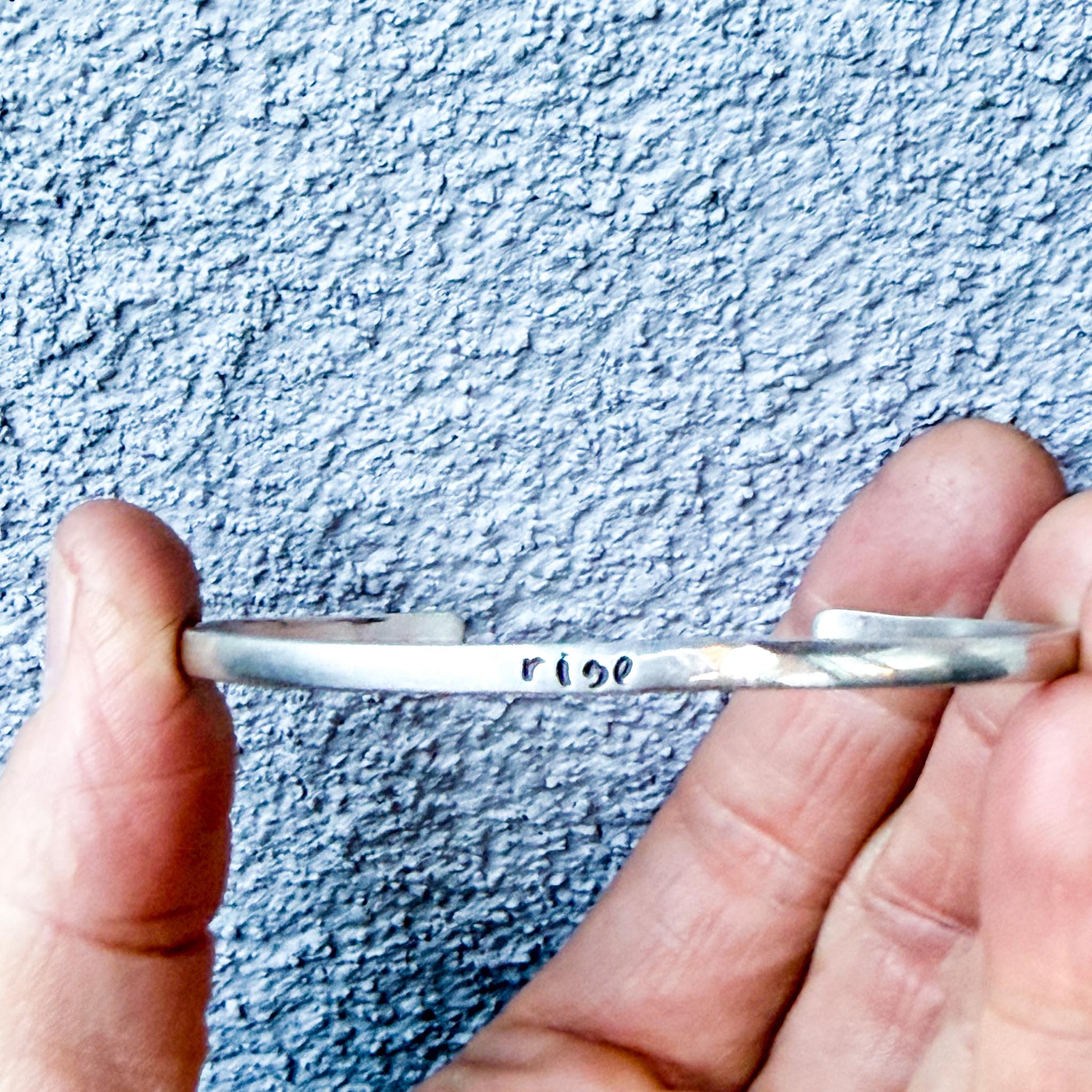 Silver bracelet held between fingers engraved text "rise" against a textured blue background