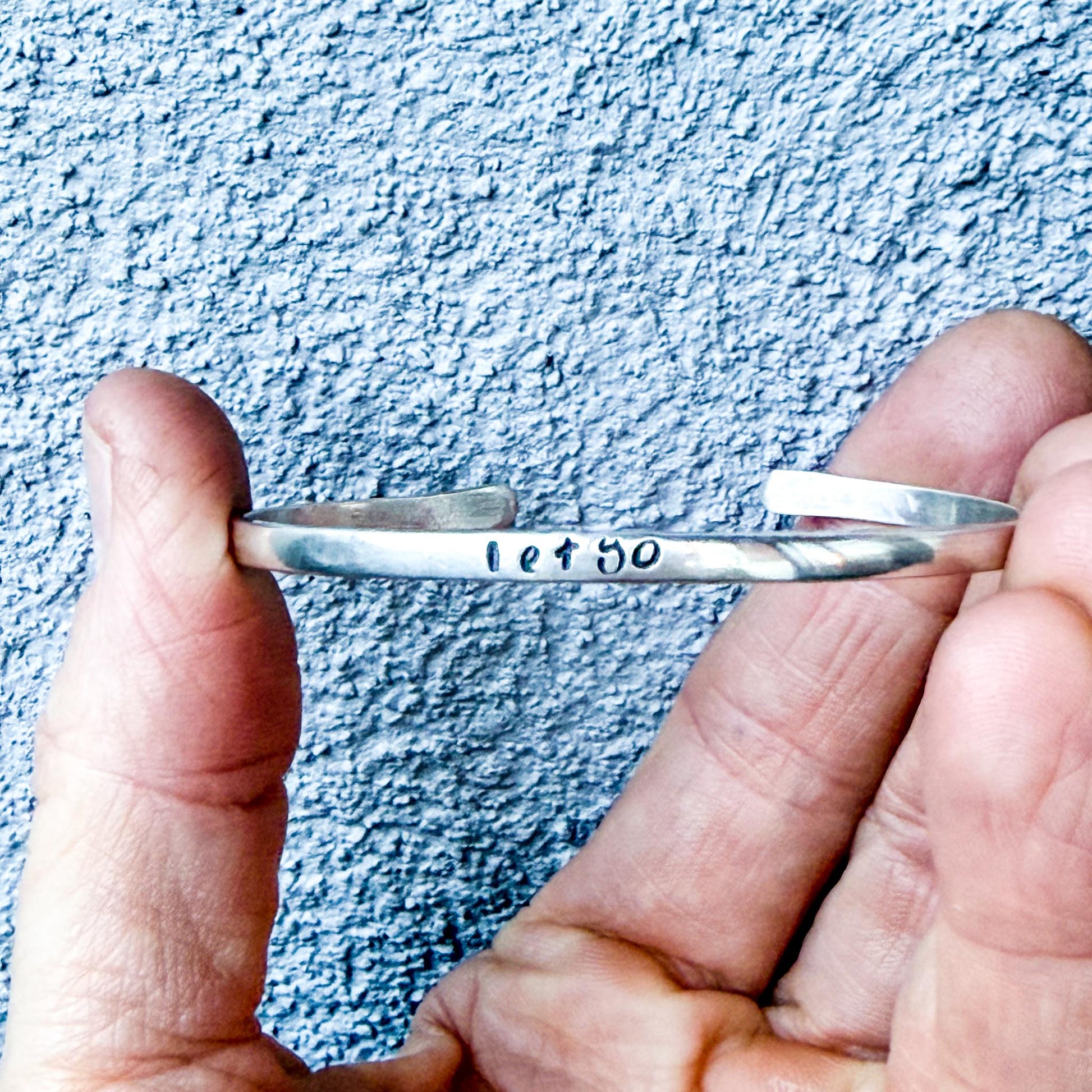 Silver bracelet with 'let go' engraving held between fingers against a textured blue background