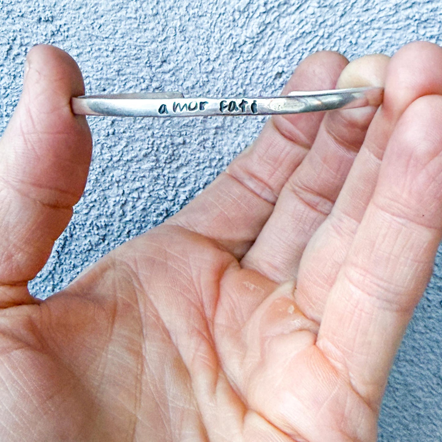 Hand holding a silver spoon with engraved text "amor fati" against a textured blue background