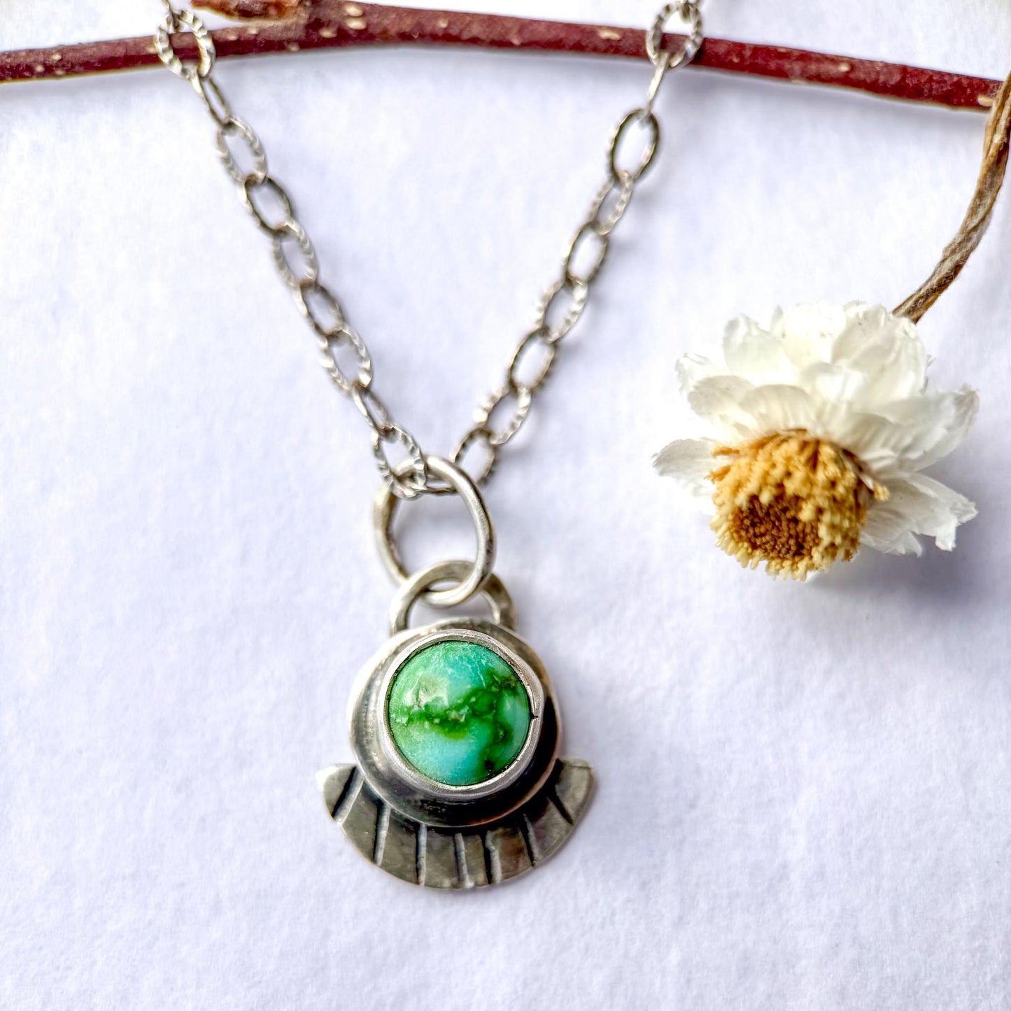 Necklace with a green pendant and chain, accompanied by a dried white flower on a white background.