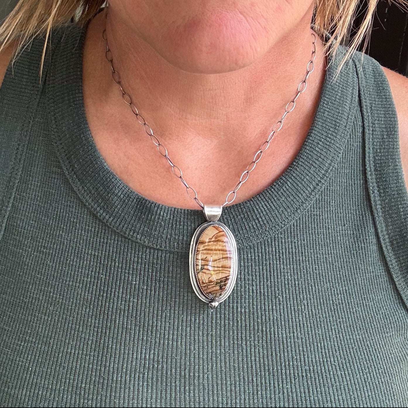 A close-up of a woman wearing a silver chain necklace with an oval pendant featuring a brown and beige wood-grain pattern, paired with a dark green ribbed tank top.