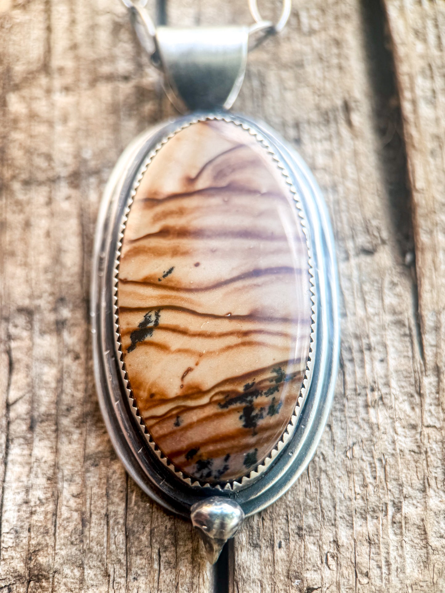 An oval pendant necklace featuring a polished brown and beige wood-grain stone in a silver bezel setting, displayed on a rustic wooden surface.