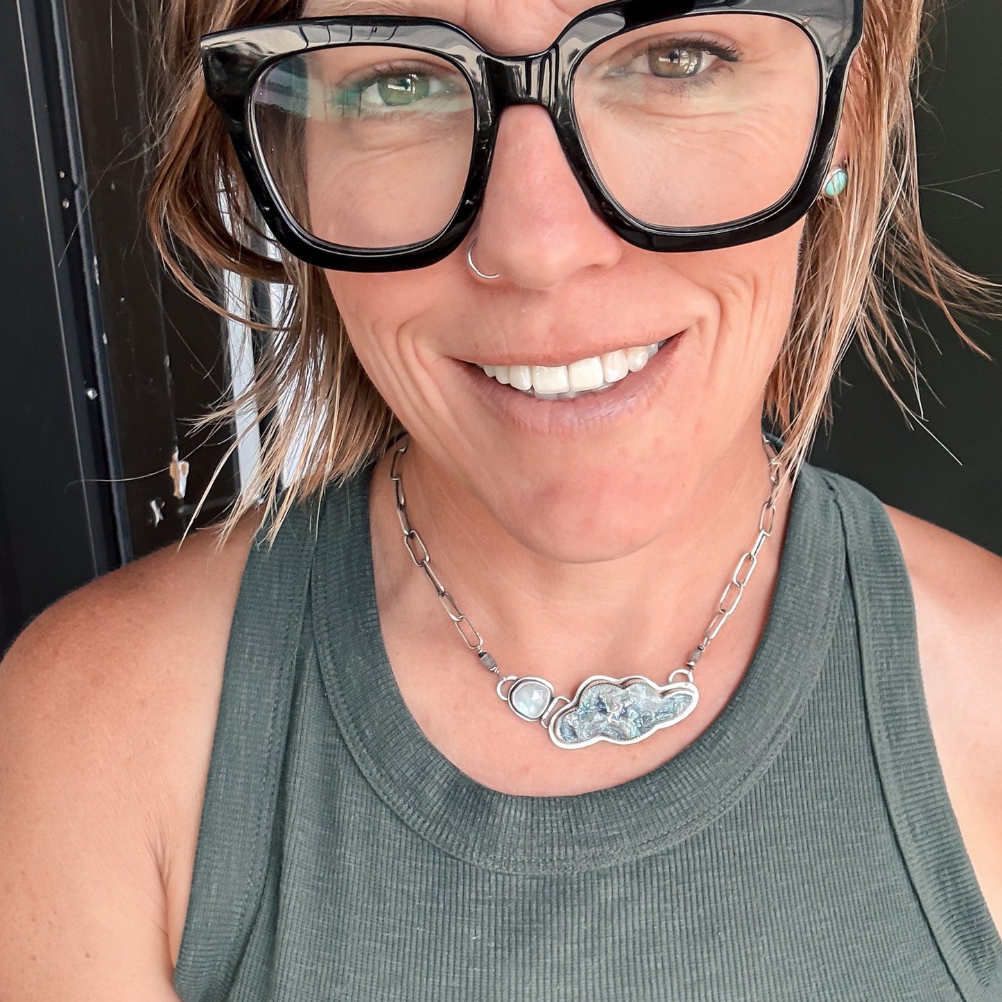 A close-up selfie of a woman wearing chunky black-framed glasses, a silver chain necklace with a polished cloud-shaped pendant, and a ribbed olive green tank top.