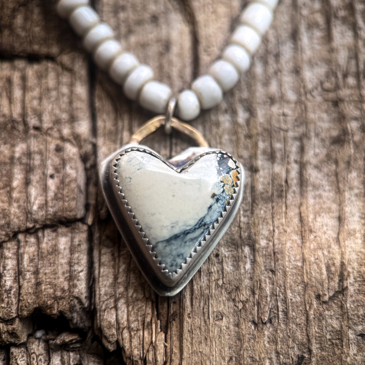 A heart-shaped pendant necklace with a white and gray marbled stone center, set in a silver-tone bezel with a gold-tone top loop, resting on a rustic wood surface. White round beads are visible at the top of the necklace.