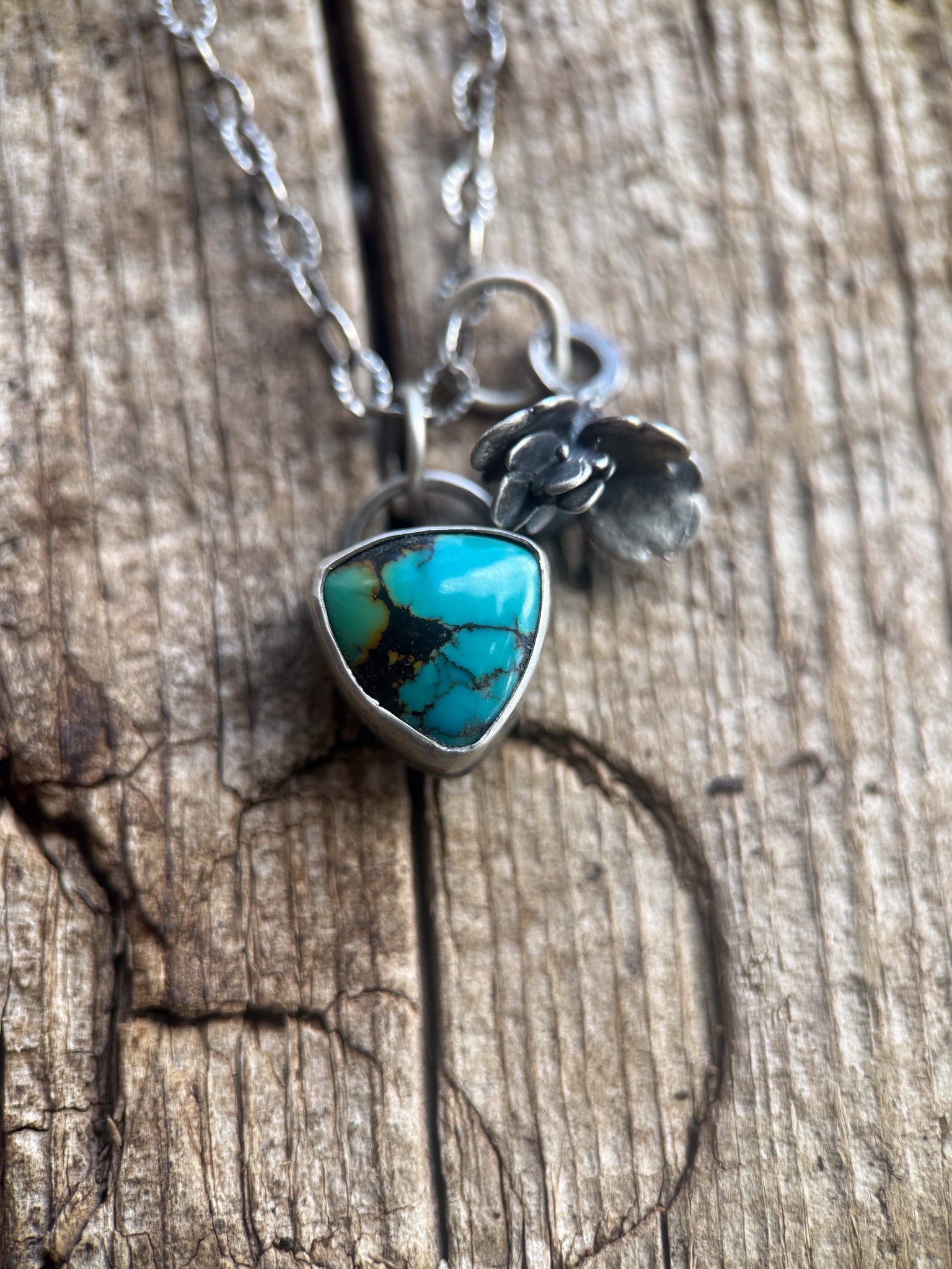 A silver necklace with a triangular turquoise stone pendant and small silver leaf charms, displayed on a rustic wooden surface.