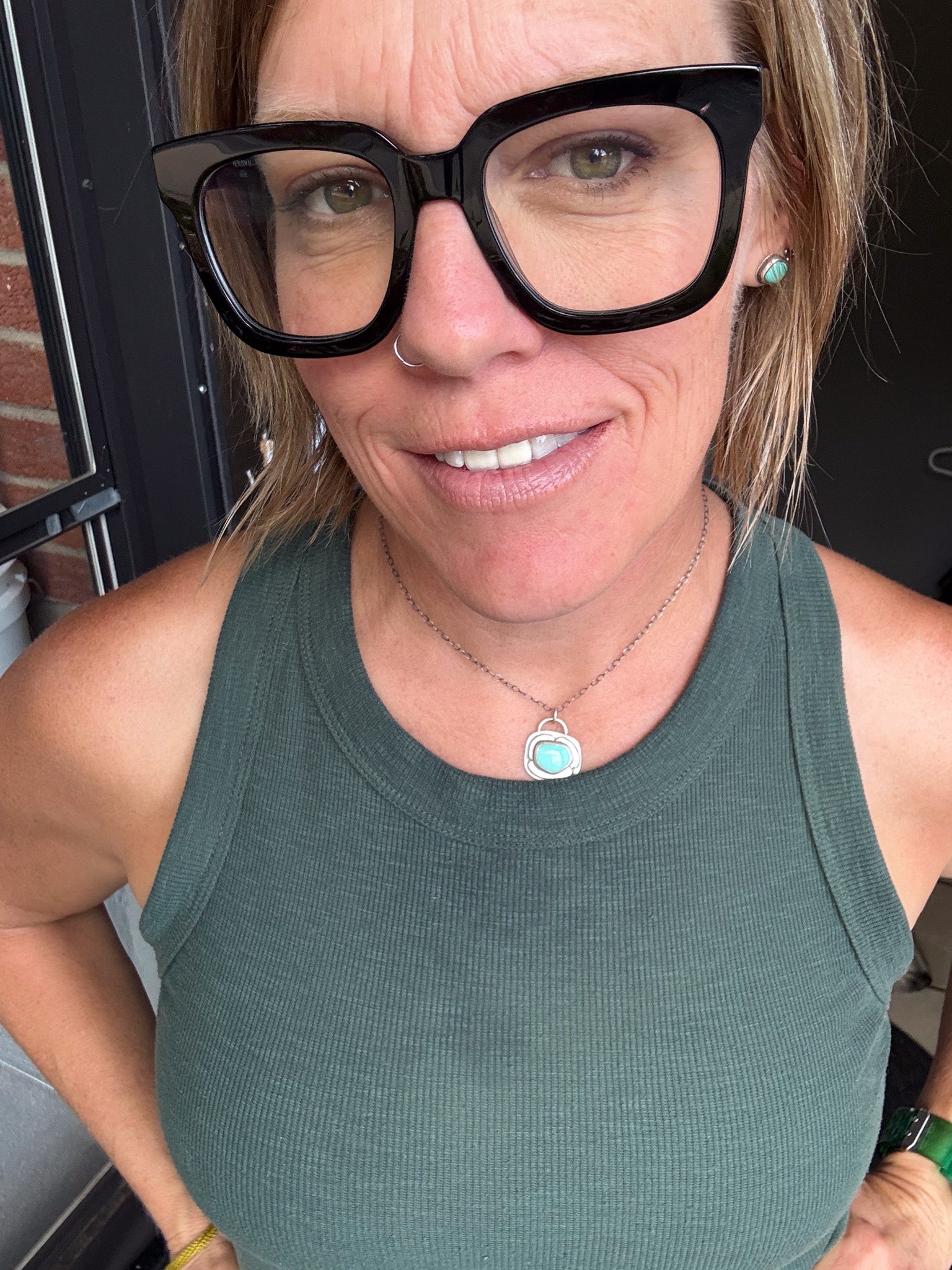 A close-up selfie of a woman wearing oversized black cat-eye eyeglasses, a sage green ribbed tank top, and a silver necklace with a turquoise pendant.
