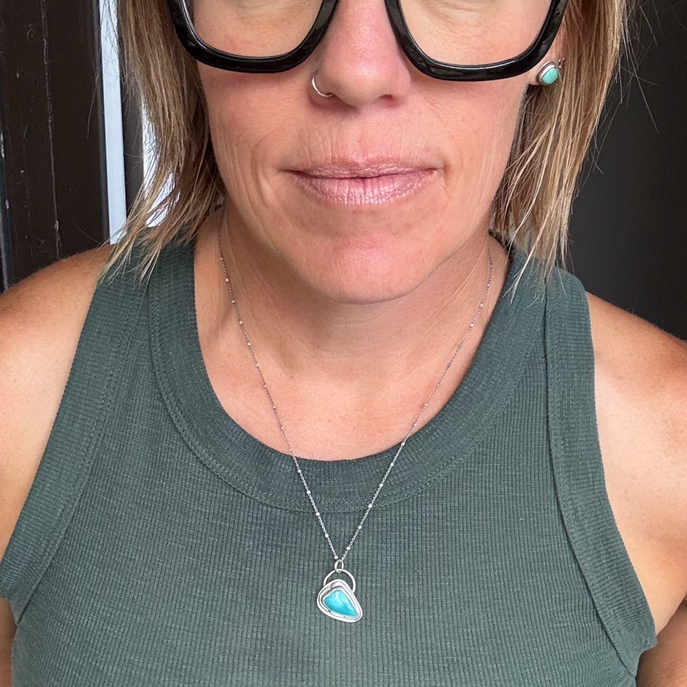 A close-up shot of a woman wearing chunky black cat-eye glasses, a silver necklace with a turquoise heart pendant, and a ribbed green tank top.