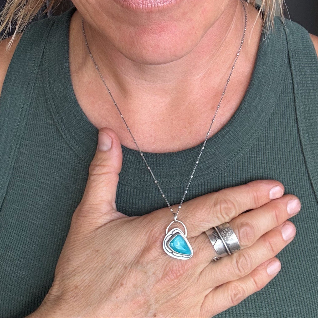 A close-up lifestyle shot of a woman wearing a silver heart-shaped pendant necklace with a turquoise blue inlay, styled with black cat-eye glasses and a green ribbed tank top.