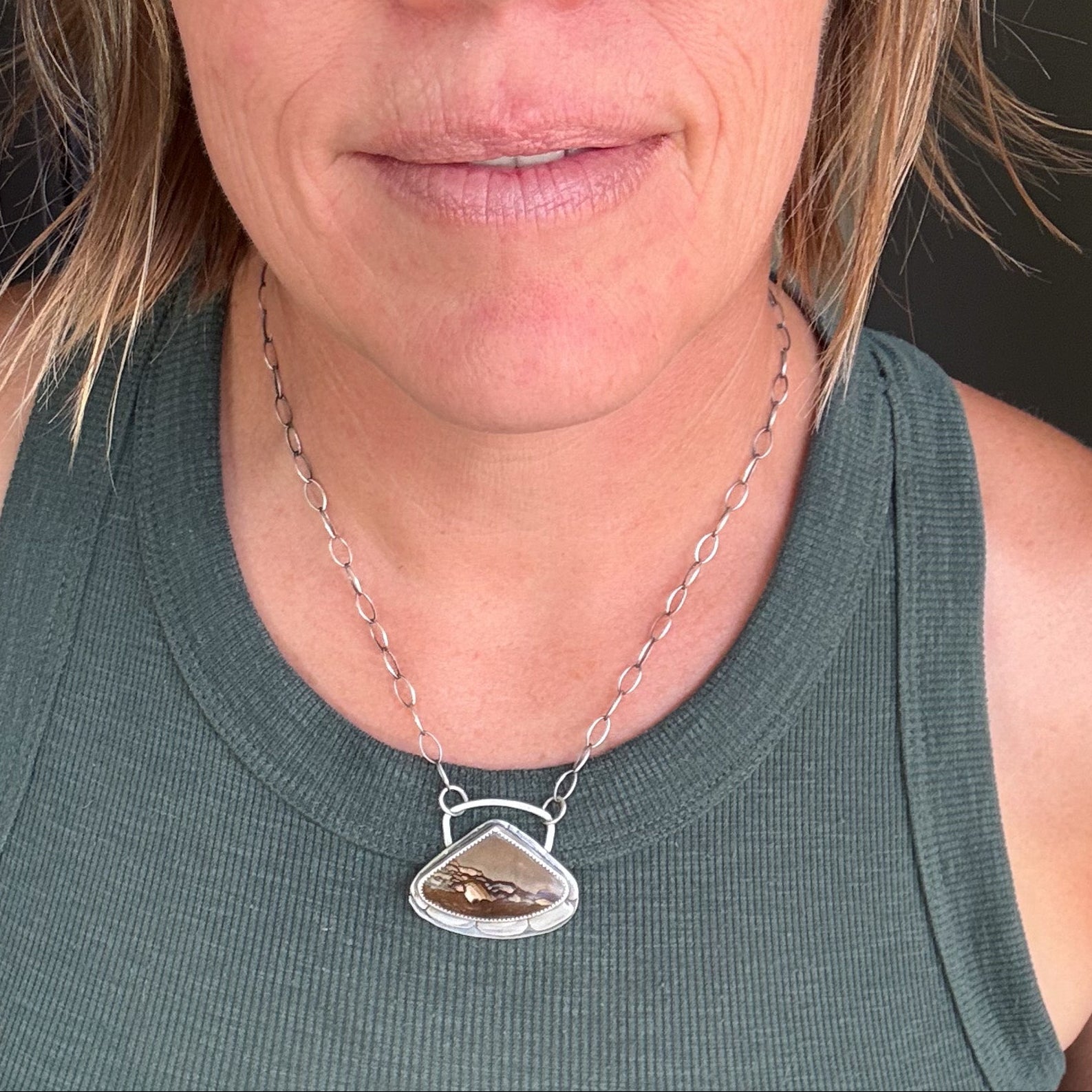 A close-up of a woman wearing a silver-toned chain necklace with a triangular pendant containing brown and gold inclusions, paired with a dark green ribbed tank top.