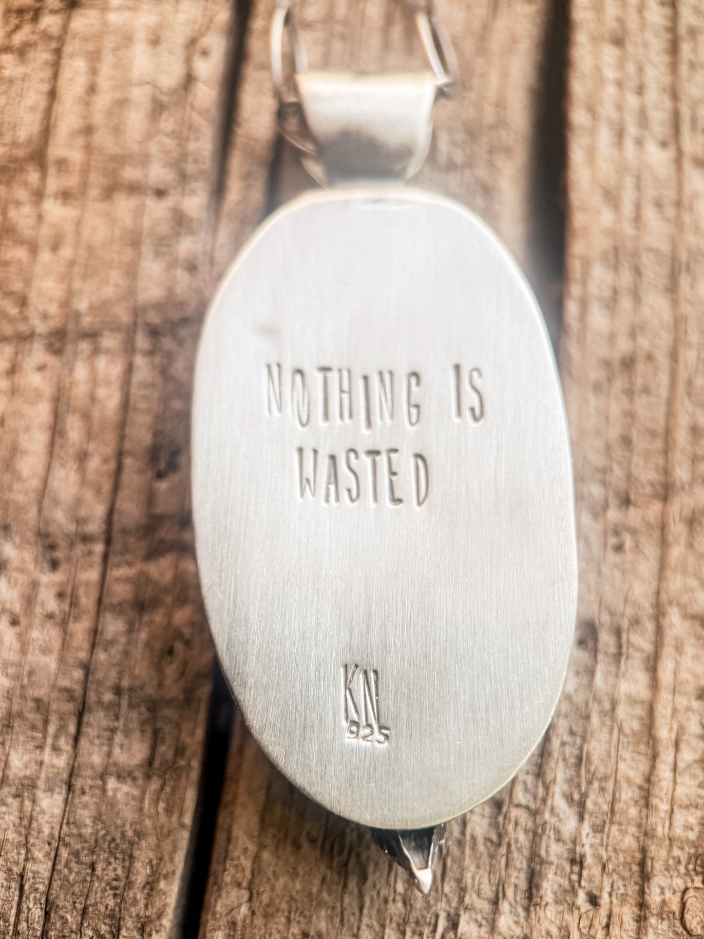 An oval sterling silver pendant with the engraved text 'NOTHING IS WASTED' and a '925' mark, resting on a rustic wooden surface.