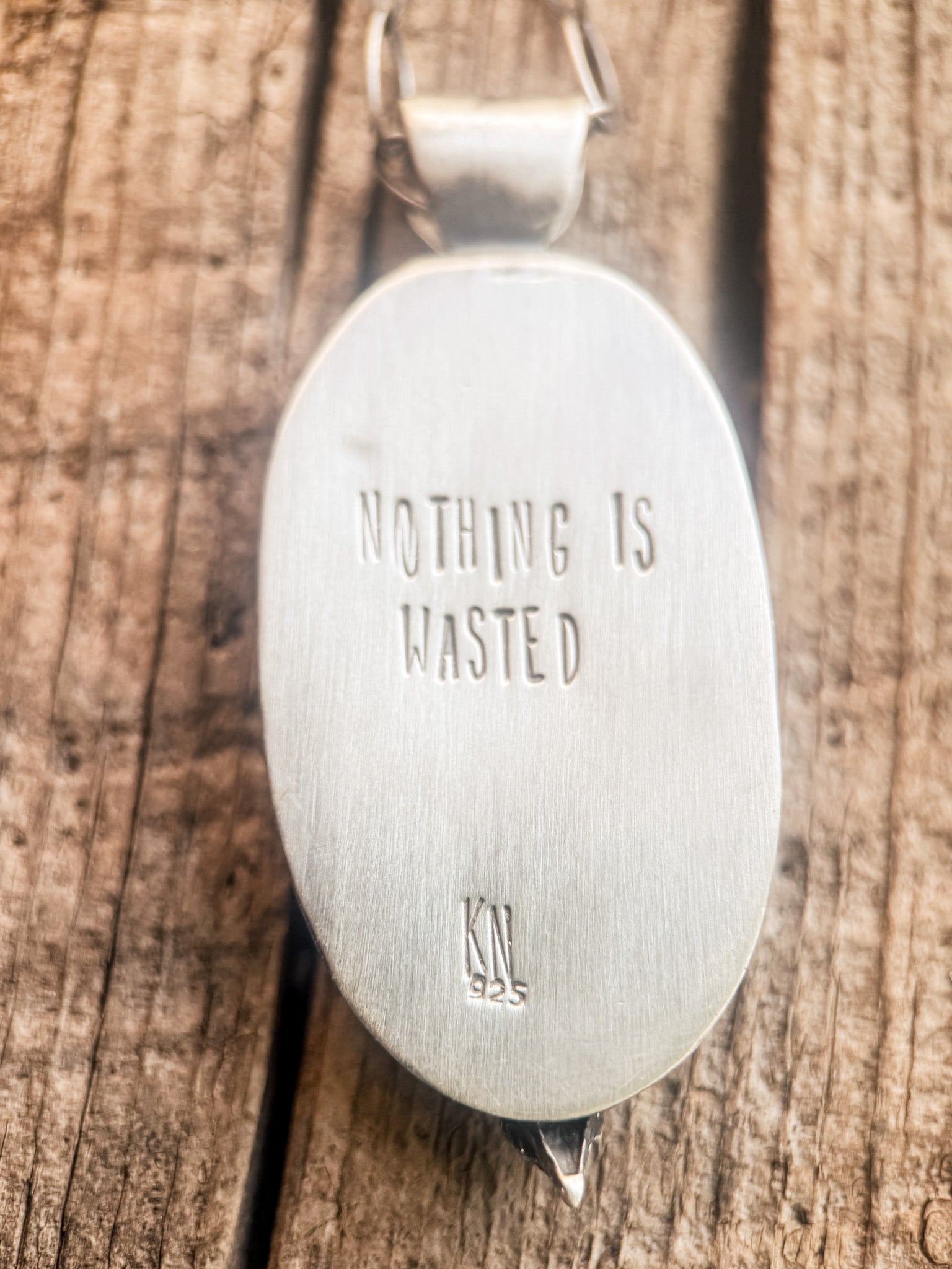 An oval sterling silver pendant with the engraved text 'NOTHING IS WASTED' and a '925' mark, resting on a rustic wooden surface.